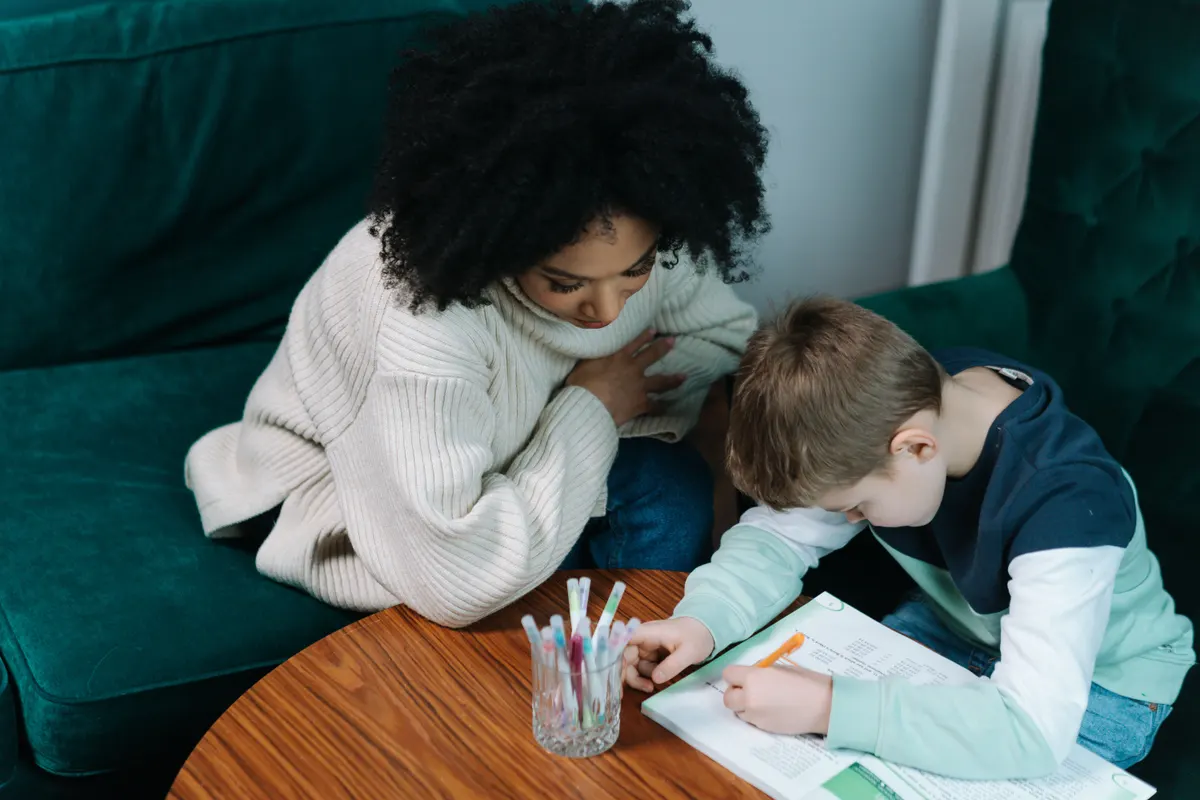 Un enfant écrit dans un cahier à la maison, sous le regard d'une personne assise à ses côtés