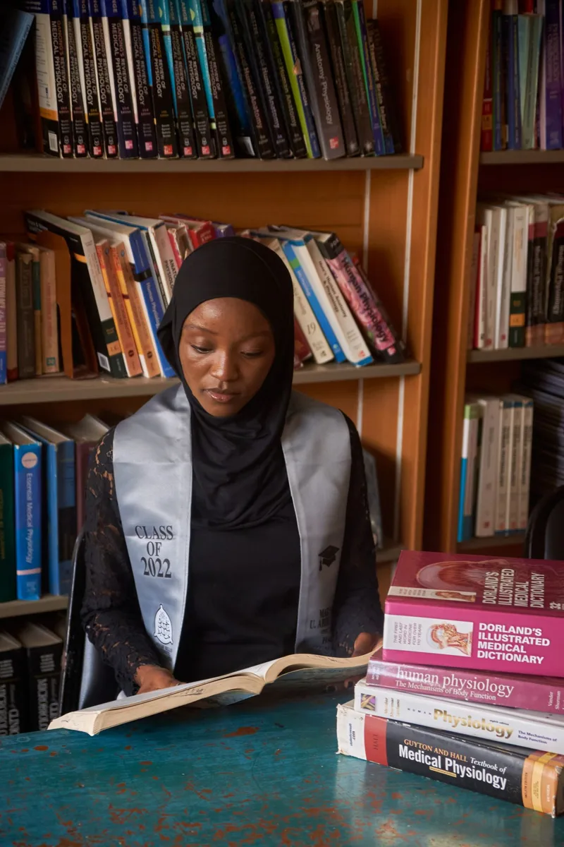 Une jeune femme en tenue de remise de diplôme lit un livre dans une bibliothèque universitaire
