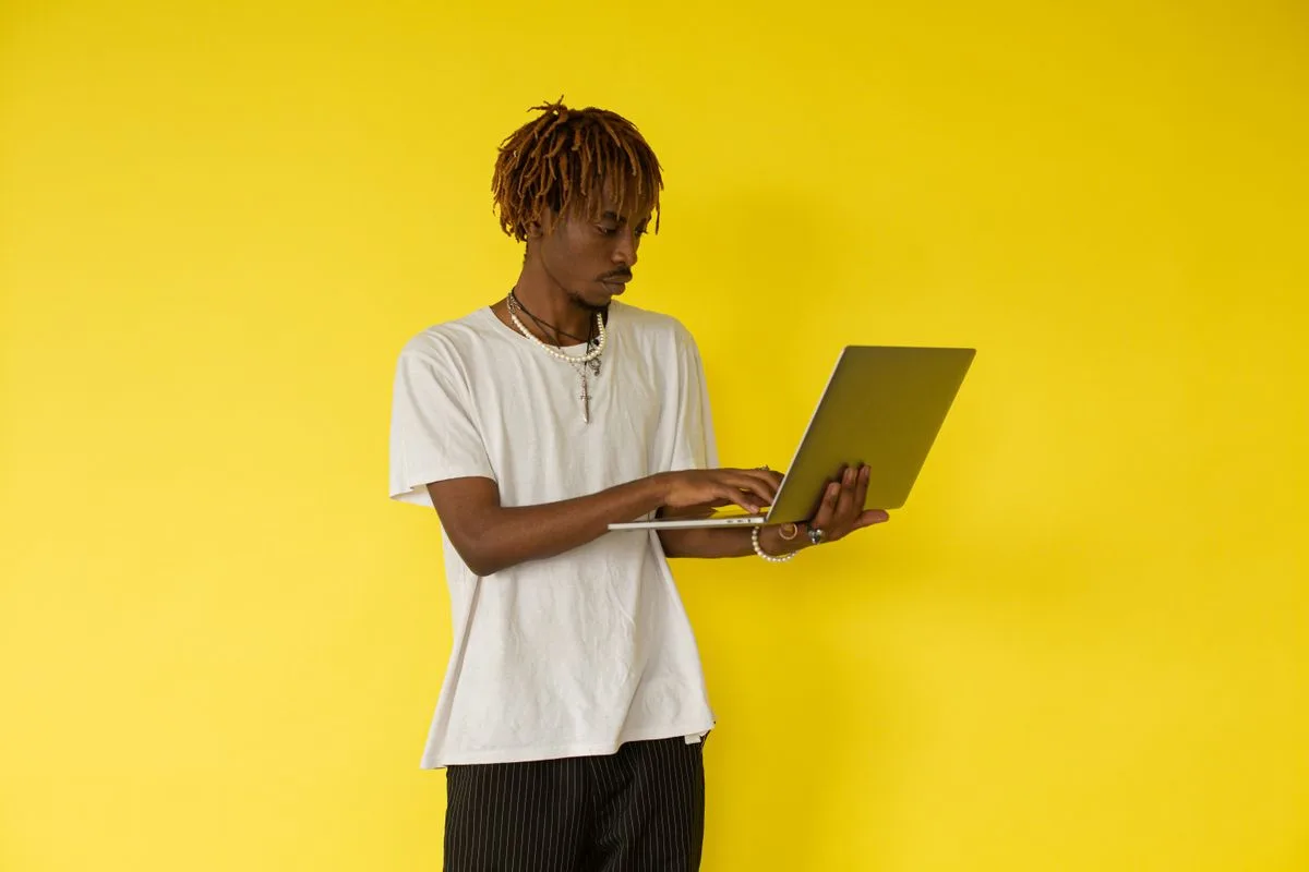 A young man stands against a plain background, typing on a laptop he holds in one hand
